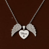 An elegant top-down studio shot of the necklace laid flat on a luxurious velvet surface, showcasing the heart with angel wings pendant, the chain length, and the engraved area, with a subtle sparkle effect on the zircon details.
